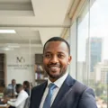 Headshot of James Karanja, a confident Kenyan male business owner in his 40s wearing an elegant navy tailored suit, smiling warmly in a sunlit modern corporate setting.
