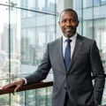 Professional portrait of David Otieno, a distinguished Kenyan real estate developer in his late 40s in a sharp grey suit against modern architectural glass.
